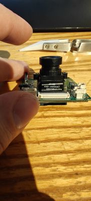 Fingers hold a small PCB with a camera module and black lens on a wooden surface.