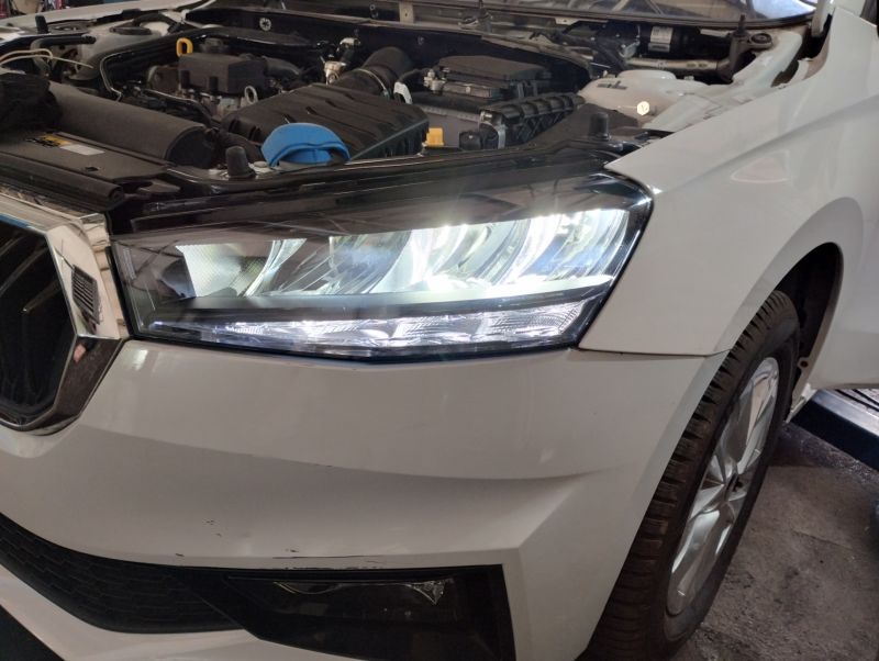Przednia lewa lampa LED włączona w białym samochodzie Skoda Fabia IV z 2023 roku, z widocznym fragmentem wnętrza komory silnika.