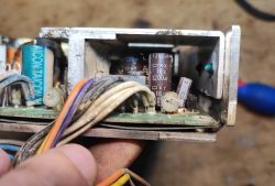 Zbliżenie na wnętrze zasilacza z widocznymi kondensatorami elektrolitycznymi, przewodami oraz śladami zabrudzenia i korozji.