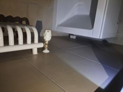 Zbliżenie na podłogę łazienki z grzejnikiem, rurką i białą skrzynką przy wannie.