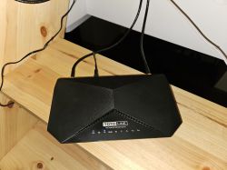Czarny router TOTOLINK stojący na drewnianej półce, z podłączonymi kablami i świecącymi diodami.