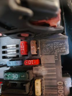 Pudełko z bezpiecznikami w samochodzie, z widocznymi kolorowymi bezpiecznikami o oznaczeniach liczbowych.