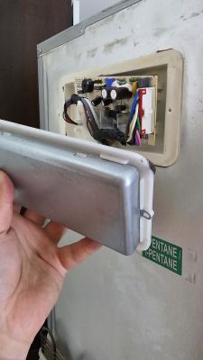 Osoba trzyma metalową pokrywę z plastikową obejmą, zdjętą z panelu z elektroniką wewnątrz urządzenia.
