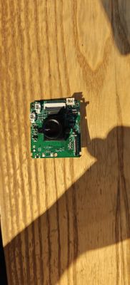 Green PCB with a camera module and protruding lens on a wooden surface in strong sunlight