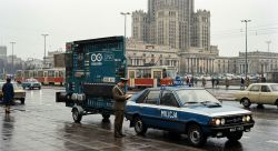 Photorealistic scene of a massive Arduino UNO board on a trailer pulled by a communist-era police car
