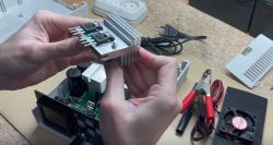 Osoba trzymająca radiator z przymocowanymi tranzystorami nad otwartym urządzeniem elektronicznym.
