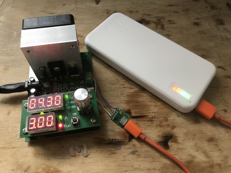 Biały powerbank z kolorowym wskaźnikiem naładowania, podłączony do testera obciążenia ze świecącymi wyświetlaczami.