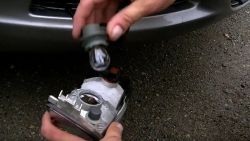 Osoba trzyma reflektor samochodowy oraz żarówkę na tle żwirowego podłoża.