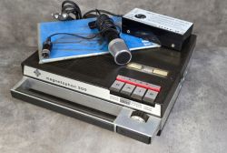 Magnetofon Uher 300 z mikrofonem, instrukcją i zasilaczem na betonowym tle