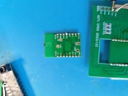 Small green PCB module with labeled pins and mounting holes, resting on a blue surface next to a larger printed circuit board.