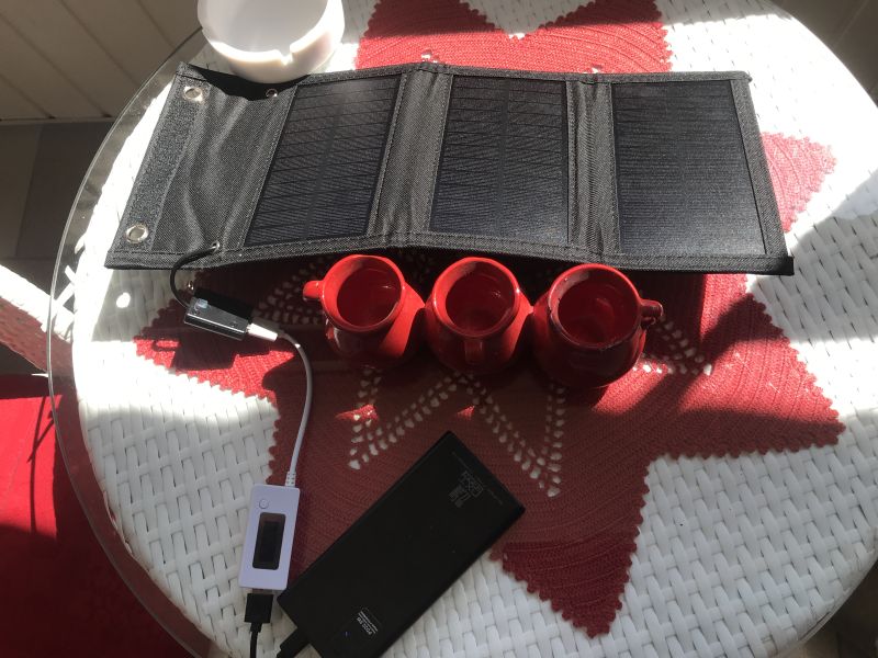 Panel solarny ładuje powerbank na balkonie, obok trzy czerwone kubki