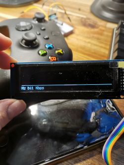 OLED display showing units Hz bit Kbps held above a gaming controller.