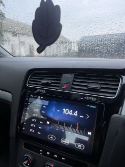 Widok wnętrza samochodu z włączonym radiem na częstotliwości 104,4 MHz i zawieszką w kształcie liścia.