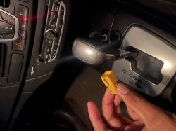 Hand holding a yellow connector in front of a car console with gear shifter and control panels.