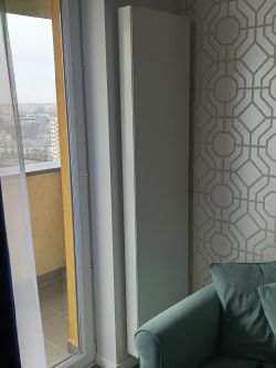 Wysoki, biały grzejnik obok drzwi balkonowych w salonie z geometryczną tapetą