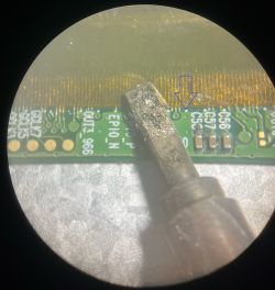 Widok zbliżony na lutownicę zbliżającą się do płytki kontrolera z zaznaczoną strzałką.