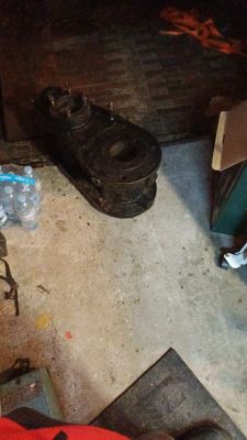 Ciężki, ciemny element metalowy na podłodze warsztatu, obok zgrzewka wody i pudełko