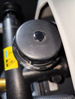 Zbliżenie na element mechaniczny z czarną plastikową pokrywką i żółtym oznaczeniem ostrzegawczym.