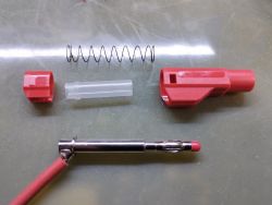 Rozłożony czerwony wtyk z elementami: sprężyna, plastikowa osłona i metalowy pin.