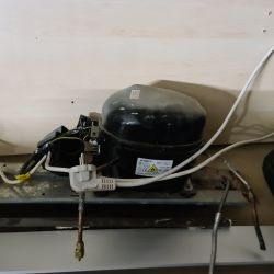 Zdjęcie czarnej sprężarki lodówkowej przytwierdzonej do metalowej podstawy, z widocznymi przewodami i wtyczką elektryczną.