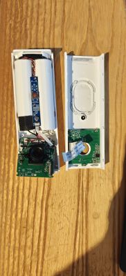 Disassembled video doorbell with housing halves, PCBs, camera lens, battery pack, and wiring on a wooden surface
