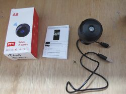 A9 IP Camera with Accessories on Table Small IP camera with box, user manual, and USB cable on a wooden surface.