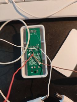 Green PCB in a white case with soldered wires on a black surface.