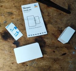 Sonoff SNZB-04 Door/Window Sensor Set on Table Sonoff SNZB-04 door/window sensor kit with manual and mounting tape on a wooden surface.