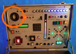 Control panel with numerous buttons, knobs, displays, and lights in a suitcase; large knob labeled “DOLACJA” in the top left, highlighted with an orange circle.