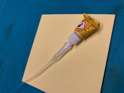Tuba kleju z nałożonym cienkim, przezroczystym wężykiem, leżąca na żółtej kartce papieru.