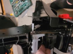 Close-up of a black mechanism with a metal spring and lever; a finger points to a part inside the housing.