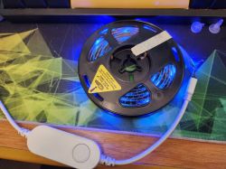 LED Strip with Controller LED strip wound on a spool with partially lit blue diodes and a connected cable, featuring a white controller with a button.