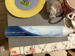 Butla CO2 z napisem „refill-co2.com” leży na stole z kolorową zastawą.