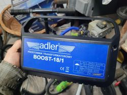 Ręka trzymająca prostownik transformatorowy Adler Boost 18/1 na tle narzędzi i sprzętu w warsztacie.