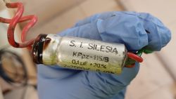 Metalowy kondensator S.I. SILESIA KPpz-115/B z pięcioma przymocowanymi przewodami, trzymany w rękawiczce.