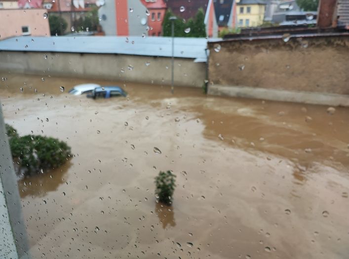 Widok zalanej ulicy z zanurzonym samochodem, widocznym przez mokre okno.