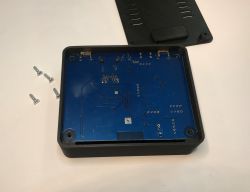 Otwarta plastikowa obudowa z niebieską płytką PCB w środku, obok leżą cztery śrubki i pokrywa.