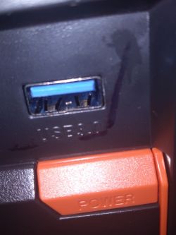 Zbliżenie na złącze USB 3.0 z metalową osłonką i odbarwieniami od markera oraz przyciskiem POWER w kolorze pomarańczowym poniżej złącza.