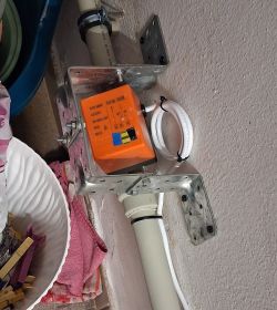 Pomarańczowy moduł elektryczny w metalowym uchwycie przy ścianie, z pętlą białego przewodu i rurą PVC