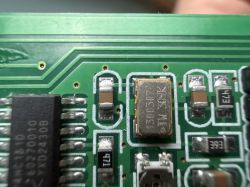Zbliżenie na zieloną płytkę PCB z oscylatorem SMD i elementami obok.
