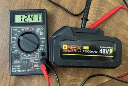 Pomiar napięcia akumulatora ONEX 48 V multimetrem Multimetr pokazuje 12,41 V obok czarnego akumulatora ONEX 48 V 15000 mAh na drewnianym blacie