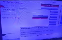 Ekran telewizora z menu „Aktualizacja oprogramowania” i paskiem postępu wyszukiwania.