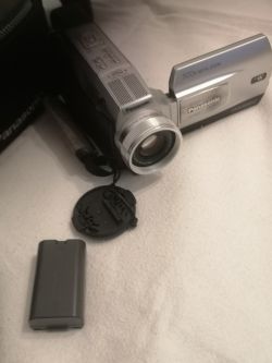 Kamera cyfrowa Panasonic z klapką obiektywu i wyjętą baterią, leżąca na jasnej powierzchni.