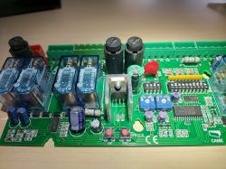 Płytka elektroniczna z różnymi komponentami, w tym przekaźnikami, kondensatorami i układami scalonymi, umieszczona na stole.