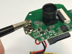 Close-up of a circuit board with a camera lens, an electronic microphone, and tweezers holding a micro USB port.