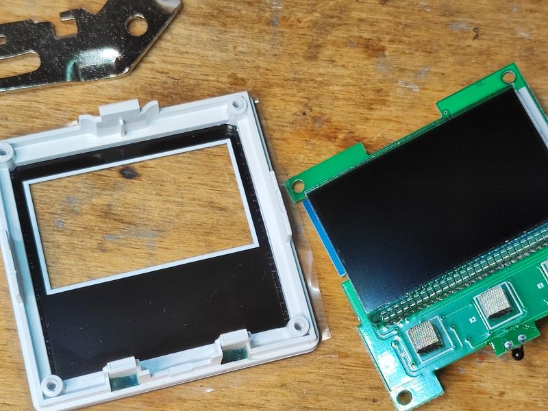 Disassembled thermostat panel: white bezel with window and a PCB with an LCD on a wooden table