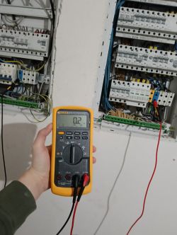 Ręka trzymająca miernik przed panelem elektrycznym.