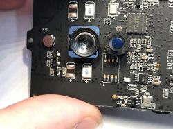 Zbliżenie na płytkę PCB z obiektywem kamery, niebieskim przyciskiem i układami SMD