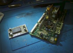 Dwie płytki PCB na niebieskiej macie serwisowej, jedna z układem scalonym, druga z wymienionymi kondensatorami.