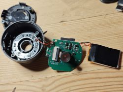 Disassembled camera showing circuit board and lens Disassembled camera with exposed PCB, lens, and battery on wooden surface